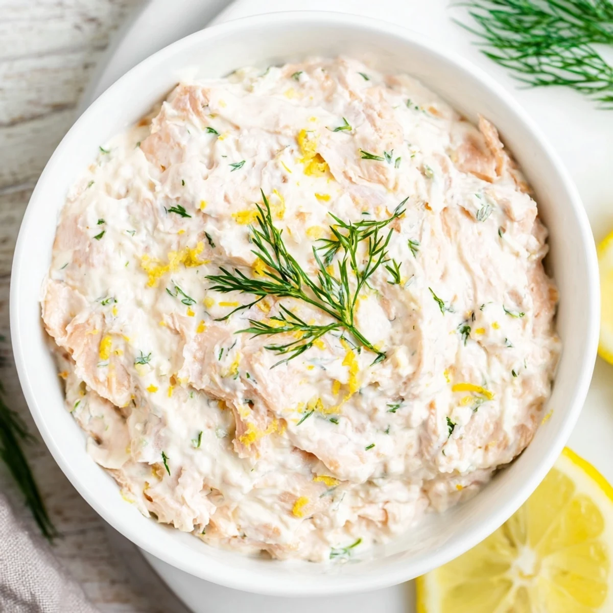 A bowl of creamy smoked salmon dip garnished with fresh dill and lemon wedges on a rustic wooden table.
